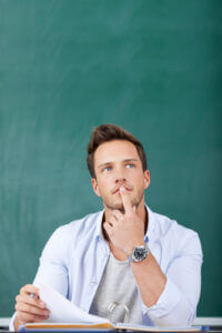 Young man sitting in front of chalkboard thinking with finger on his chin