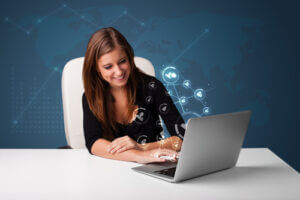 Young lady sitting at desk and typing on laptop with social network icons coming out
