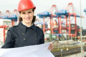 Female civil engineer wearing helmet and checking the drawings in front of industry harbor background 