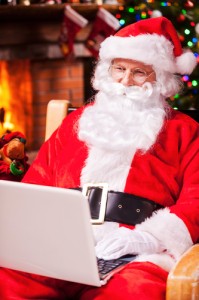 Modern Santa Claus. Cheerful Santa Claus working on laptop and smiling while sitting at his chair with fireplace and Christmas Tree in the background 